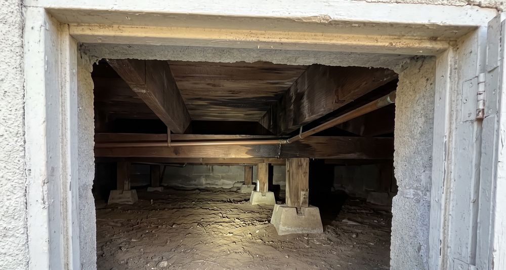 A low crawl space seen through an access opening in a concrete foundation wall, with floor joists, pier blocks and a copper water line